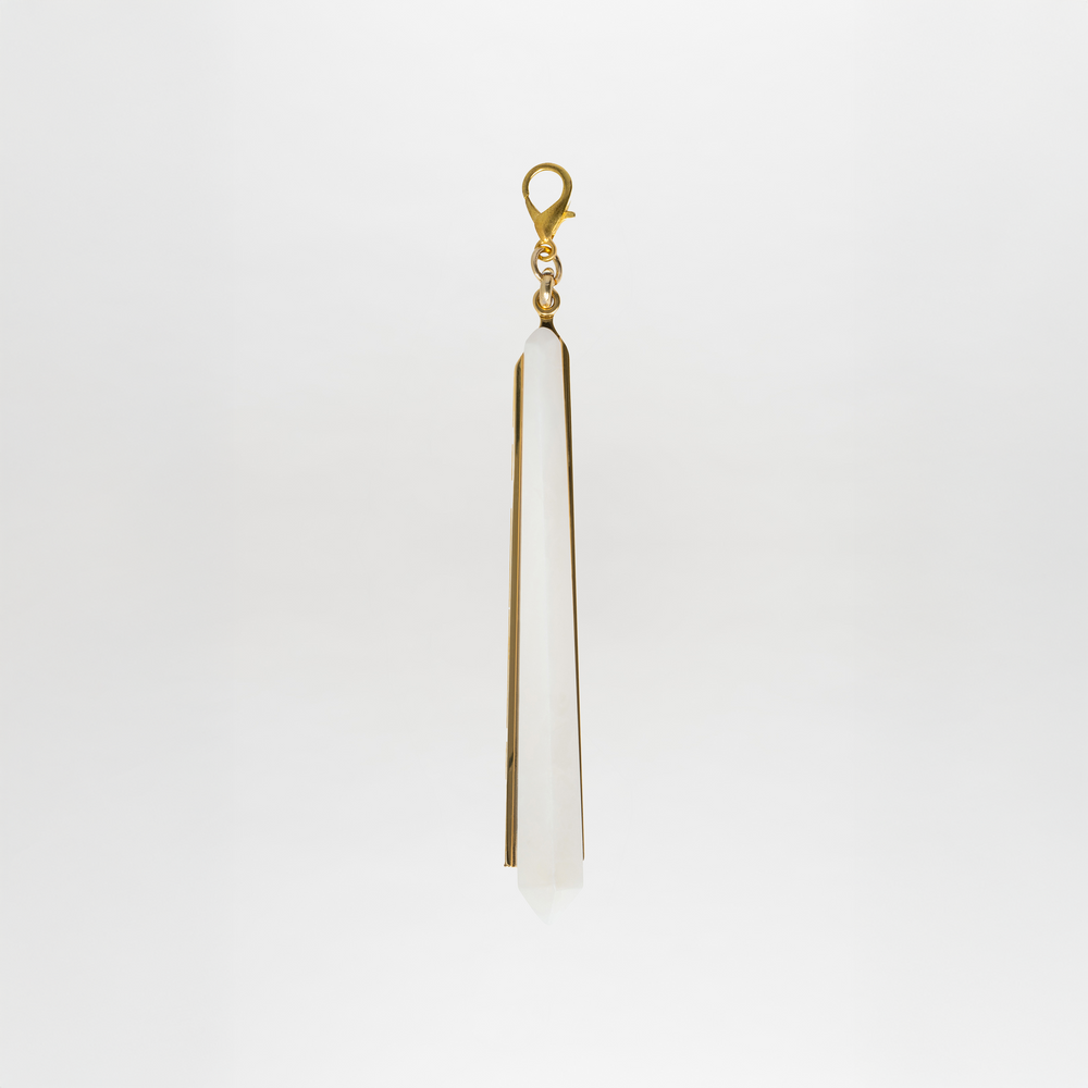 The Montse Charm features a long, pointed crystal pendant with a gold-plated brass clasp, set against a plain white background.