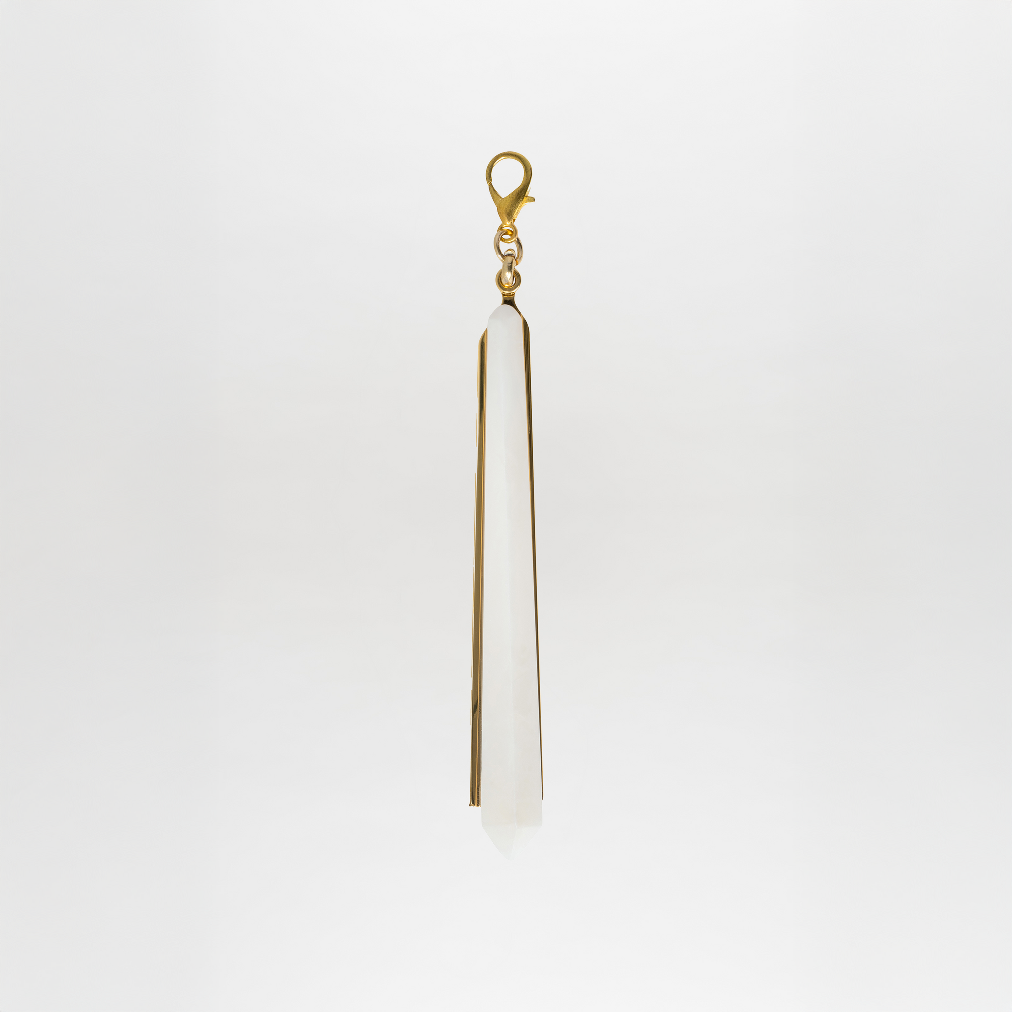The Montse Charm features a long, pointed crystal pendant with a gold-plated brass clasp, set against a plain white background.