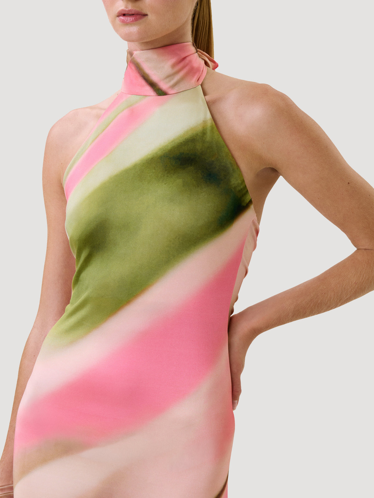 A person wearing the Maika Dress—a sleeveless halter-neck style featuring pink, green, and cream diagonal patterns—stands with one hand on their hip against a plain background.