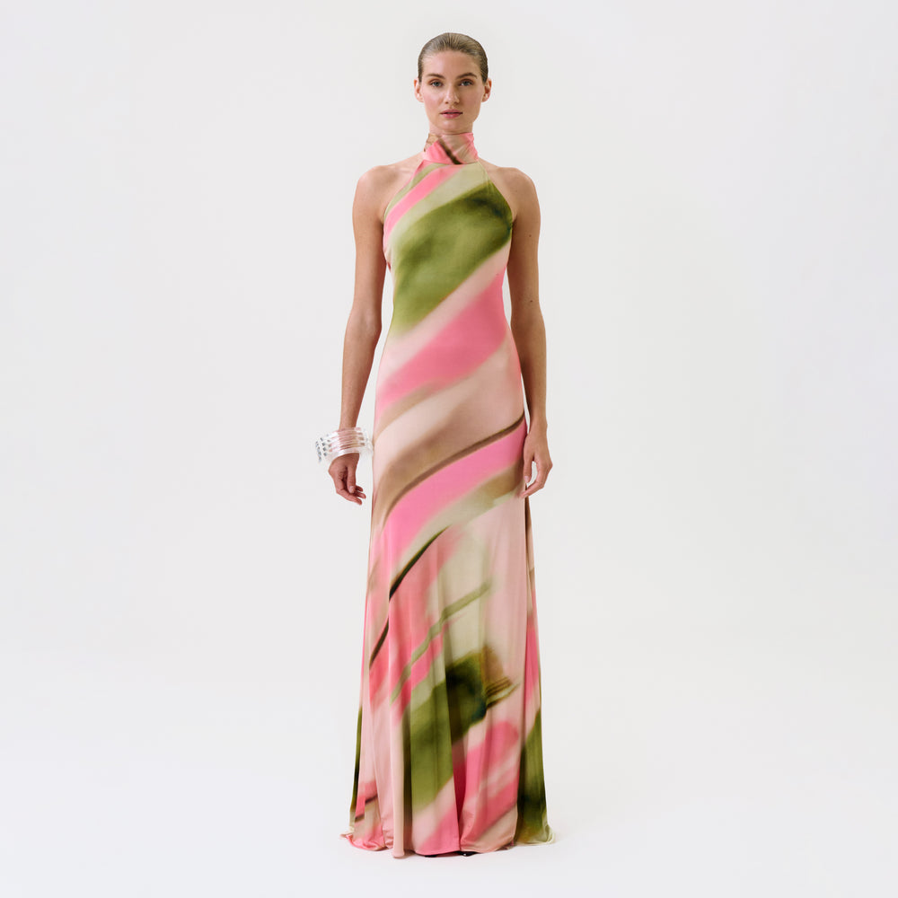 A woman stands against a white background, wearing the Maika Dress—a long, halter-neck dress featuring a pink and green abstract pattern—paired with a chunky white bracelet.