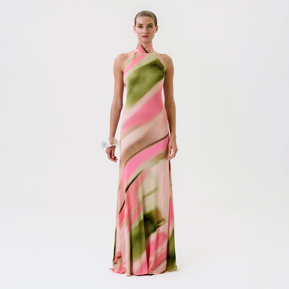 A woman stands against a white background, wearing the Maika Dress—a long, halter-neck dress featuring a pink and green abstract pattern—paired with a chunky white bracelet.