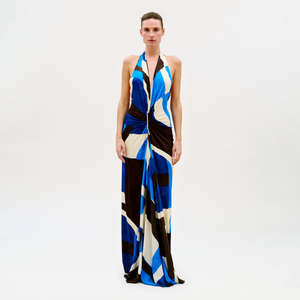 A person poses against a plain light background wearing the Malek Dress, a long halter-neck style with a bold blue, black, white, and beige geometric pattern and a knotted waist detail.