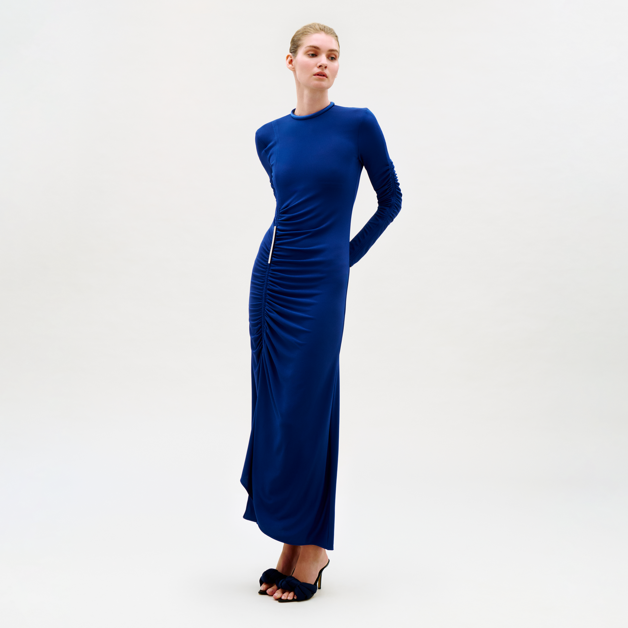A woman stands against a plain background wearing the floor-length, long-sleeved Mara Dress in royal blue with ruched waist and center details, paired with black open-toe heels. Her hair is pulled back, and her hands are behind her back.