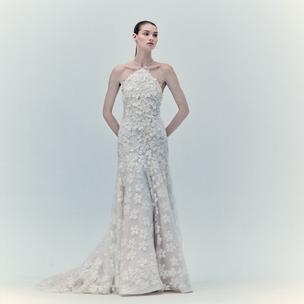A woman wearing the Maria Dress—a floor-length white maxi with a halter neck and delicate floral appliqué—stands with her hands in her pockets against a pale, minimalist background.