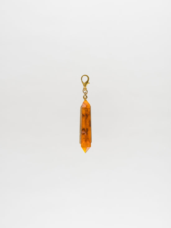 Presenting the Mariela Charm Amber: a pendant featuring an orange crystal with a metal clasp, showcasing a Petralux® fantasy stone against a plain white background.