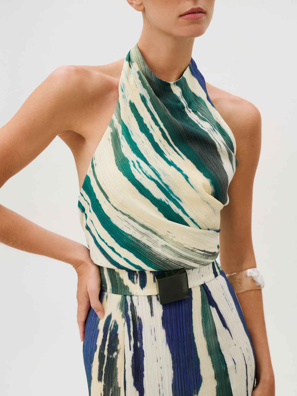 The Marwa Dress is a crepe de viscose maxi dress featuring a sleeveless, halter-neck silhouette with a pleated, abstract green, blue, and cream brushstroke print and a black rectangular belt buckle at the waist.