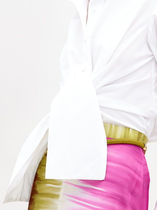 A person wearing a Mercury Blouse White and a vibrant pink skirt with green accents, standing against a plain white background, available for pre-order now. Ships by November 15th, 2024.