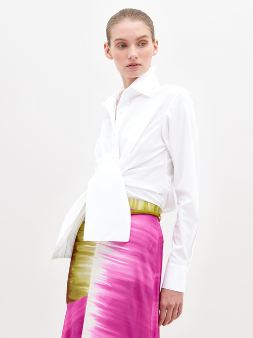 A person wearing a Mercury Blouse White and a vibrant pink skirt with green accents, standing against a plain white background, available for pre-order now. Ships by November 15th, 2024.