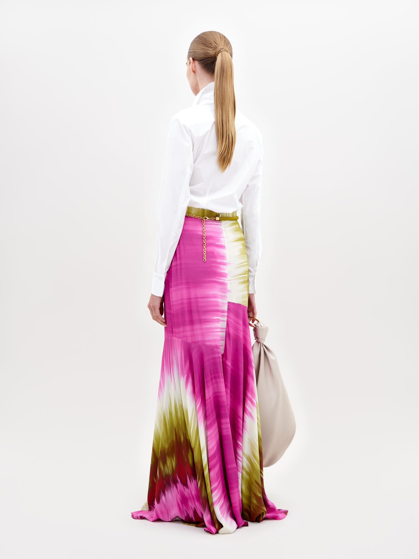 A person stands wearing a white collared shirt tucked into a long, flowing Shane Skirt Violeta Lime Tie Dye with patterns in pink, green, and white. The background is plain and light-colored. Secure this stylish look by placing a pre-order now to ensure it arrives before the ship by date of November 15th, 2024.