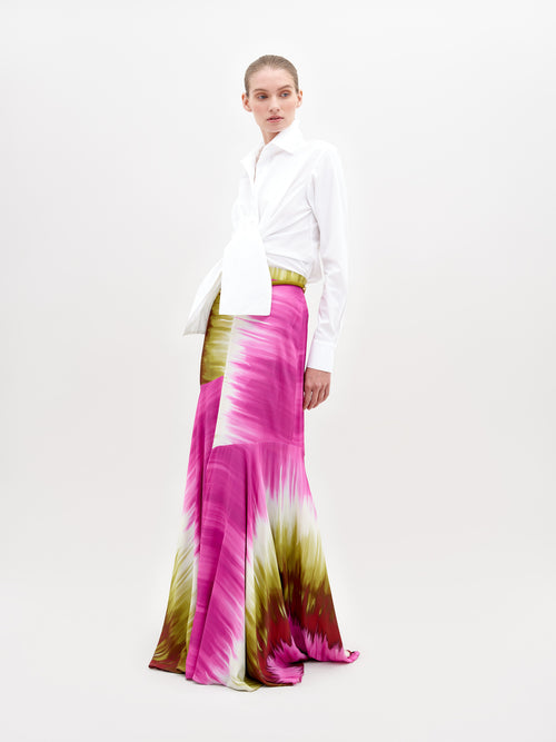 A person stands wearing a white collared shirt tucked into a long, flowing Shane Skirt Violeta Lime Tie Dye with patterns in pink, green, and white. The background is plain and light-colored. Secure this stylish look by placing a pre-order now to ensure it arrives before the ship by date of November 15th, 2024.