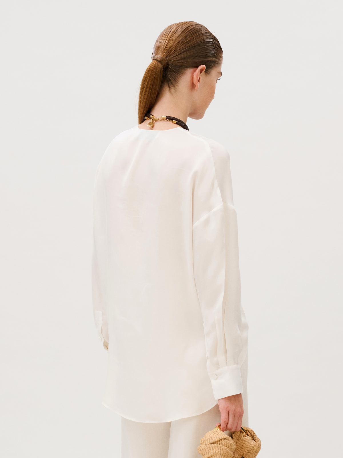 A person seen from behind wearing an off-white long-sleeved Merida Blouse and carrying a woven tan handbag.
