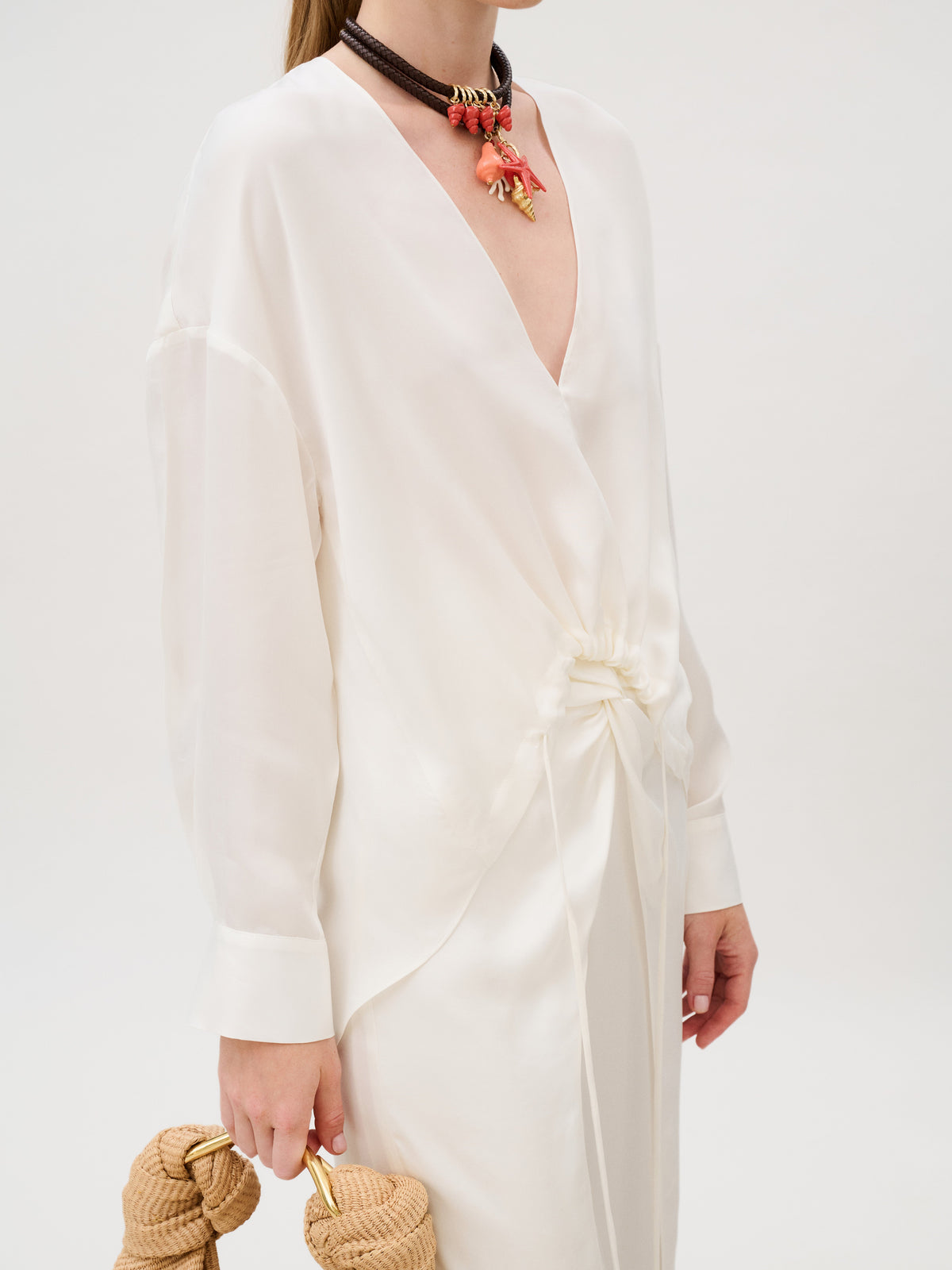The Merida Blouse is a long-sleeved white Cupro top with a deep V-neck and cinched front detail, styled with a layered beaded necklace featuring coral accents and a woven straw handbag with gold handles.