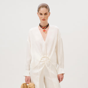 A person wearing a white, long-sleeved Merida Blouse in cupro with a V-neck, a gathered drawstring waist, and a layered beaded necklace featuring a starfish charm.