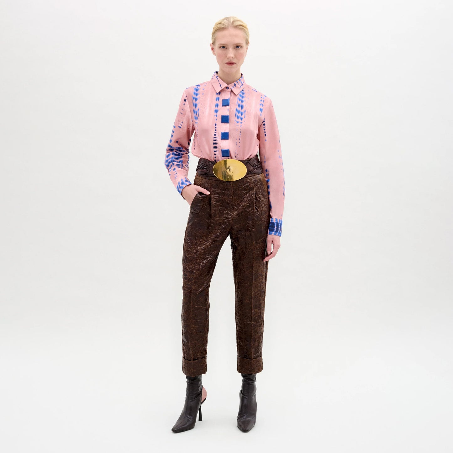 A person stands against a plain white background wearing the Moad Pant, a high-waisted, straight-leg, cropped style in Italian fabric, styled with a pink patterned shirt, large gold belt, and pointed dark ankle boots. One hand is in their pocket.