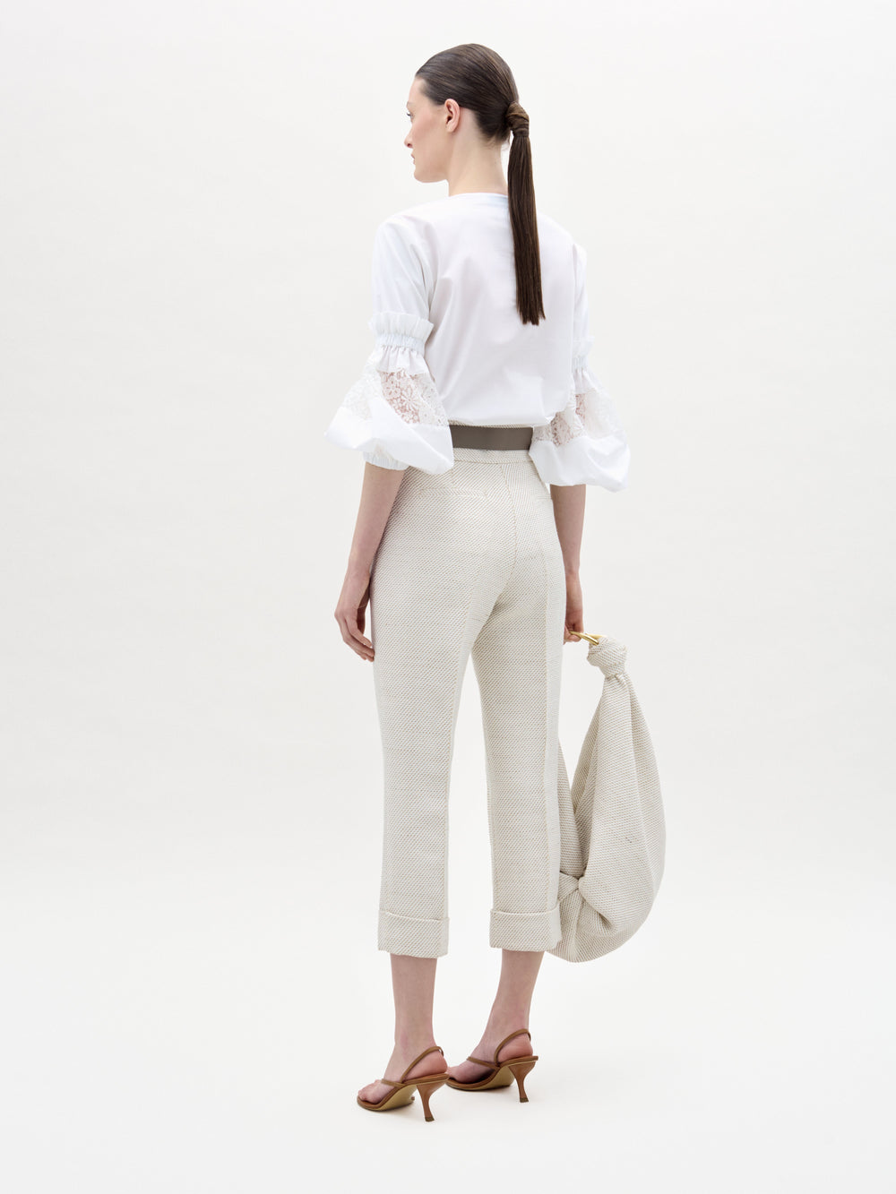 Woman in a white blouse and beige pants holding a beige bag on a white background