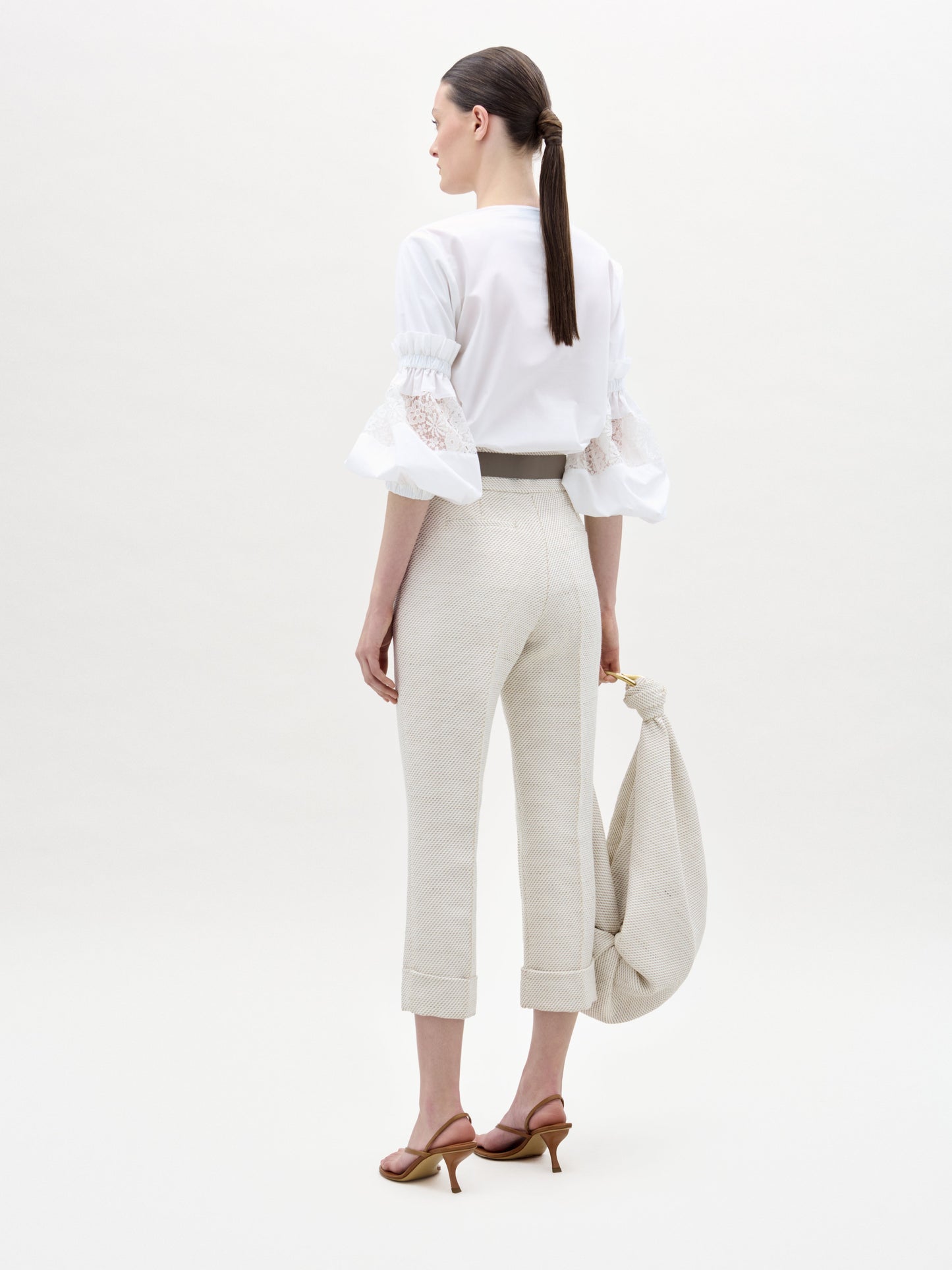 Woman in a white blouse and beige pants holding a beige bag on a white background