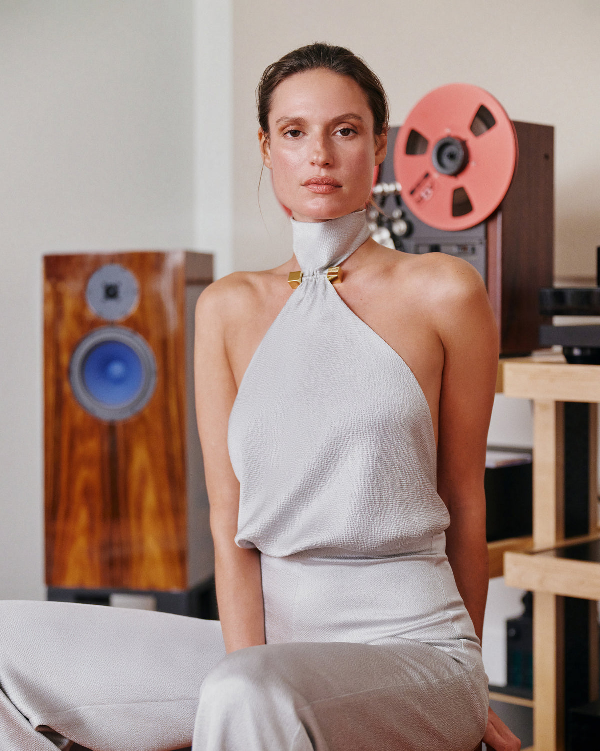 A woman wearing a light gray halter-neck top and matching pants sits in a room with vintage audio equipment, including speakers and a reel-to-reel recorder, in the background.