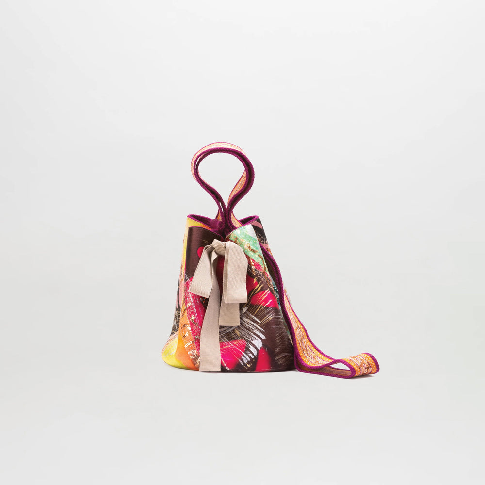 The Mochila Laaput Small is a vibrant, multicolored bag with a unique twisted handle, central decorative bow, and sculptural shape—beautifully handcrafted in Colombia and shown against a plain white background.