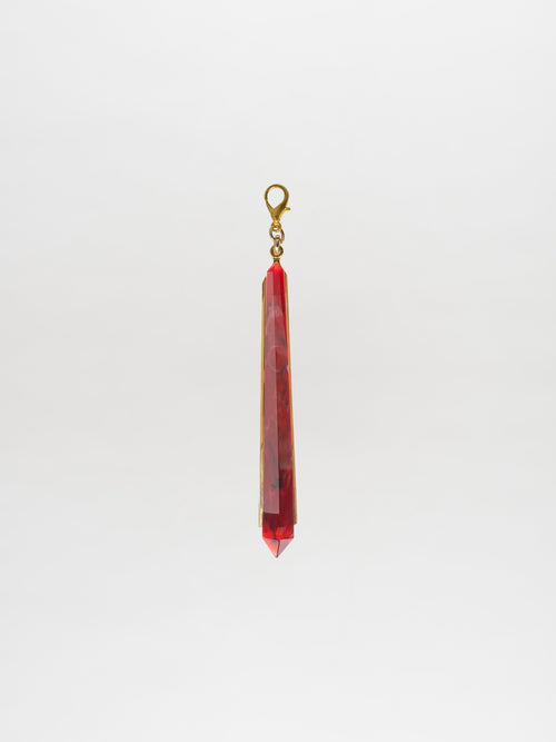 The Montse Charm Rouge, a geometric pendant featuring a red crystal and a gold-plated brass clasp, is elegantly displayed against a plain white background.