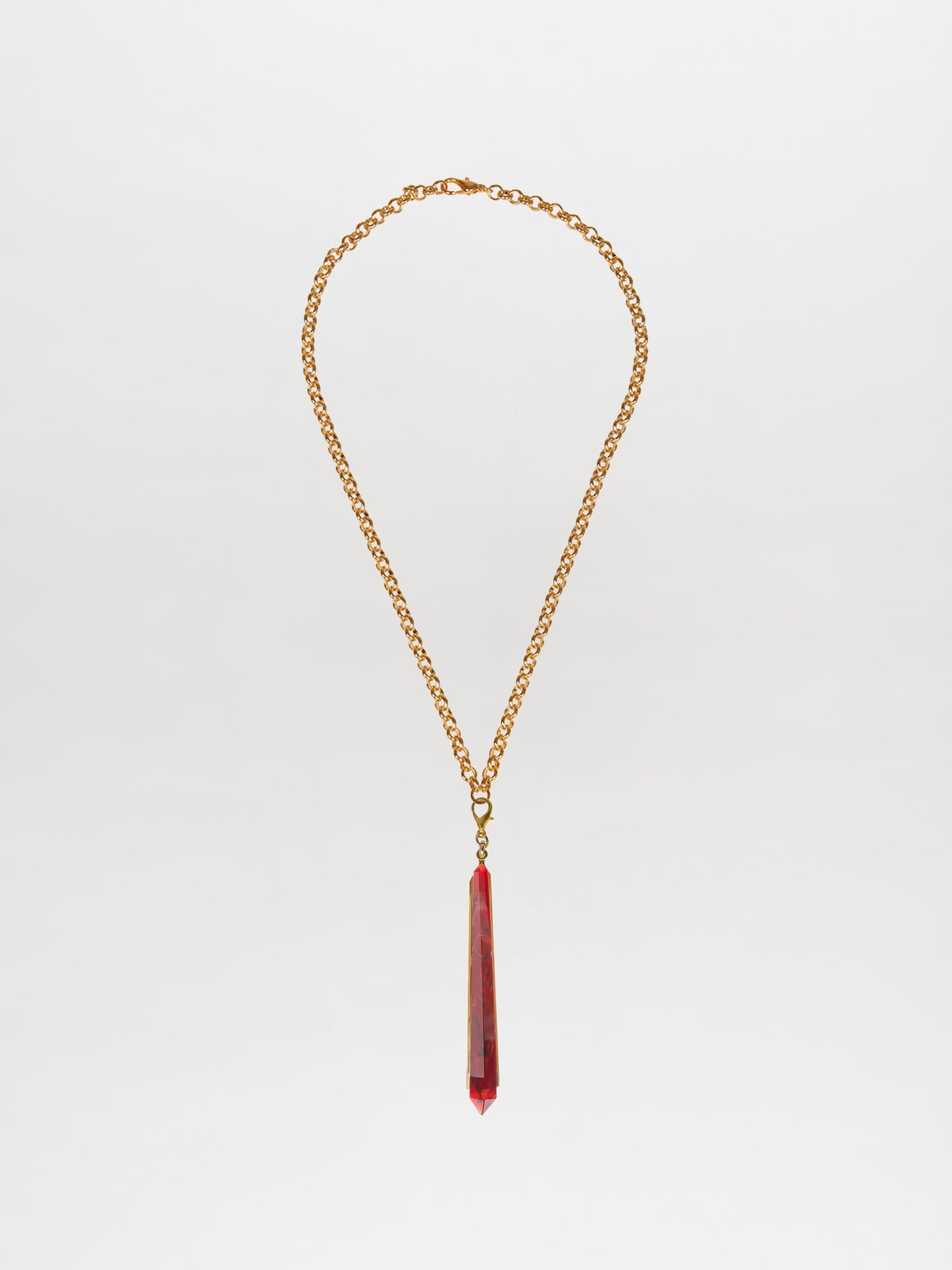 The Montse Necklace Rouge is a gold chain necklace showcasing an elongated, geometric crystal pendant in vivid red, set against a plain white backdrop. Ideal for layering, this piece adds a touch of elegance to any outfit.