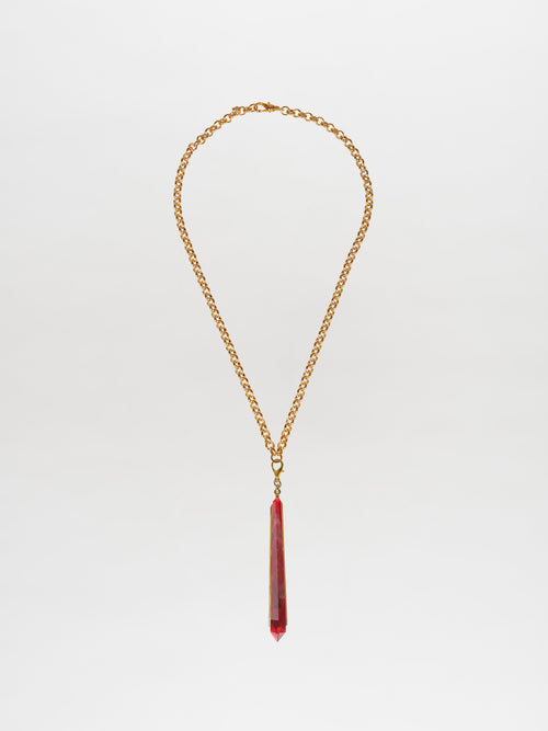 The Montse Necklace Rouge is a gold chain necklace showcasing an elongated, geometric crystal pendant in vivid red, set against a plain white backdrop. Ideal for layering, this piece adds a touch of elegance to any outfit.