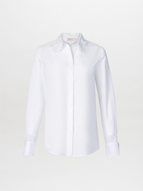 The Naroa Blouse White is a cotton button-up with a collar, featuring floral embroidery on the shoulders and lace fringe detailing, set against a plain background.