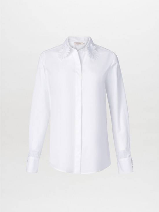 The Naroa Blouse White is a cotton button-up with a collar, featuring floral embroidery on the shoulders and lace fringe detailing, set against a plain background.