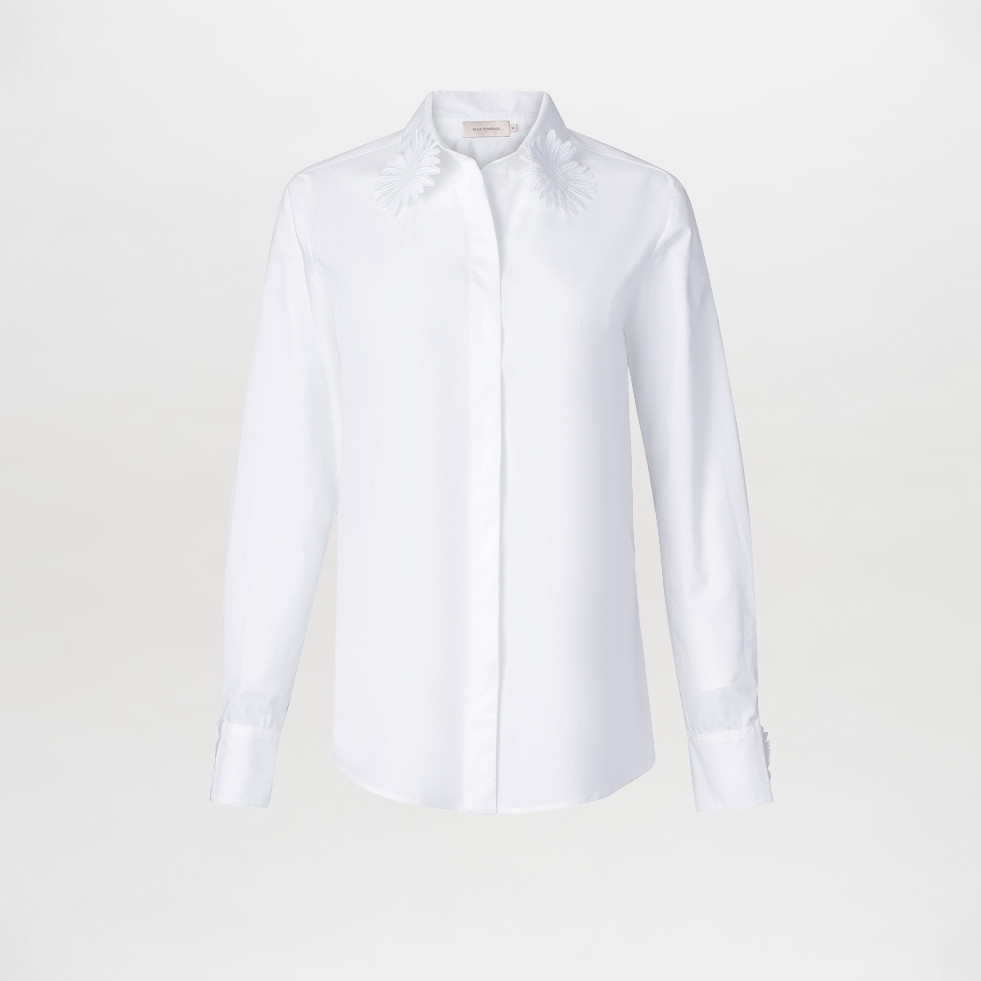 The Naroa Blouse is a white cotton top with long sleeves, a classic collar, and delicate embroidered floral details on the collar tips. Made in Colombia, it’s shown against a plain white background.