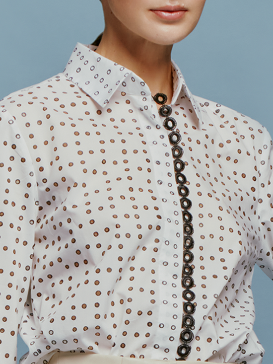 A woman stands against a blue background, wearing the white Naroa Blouse with black dots and light gray pants, accessorized with a chain belt, ready for the fresh styles of September 2nd, 2024.