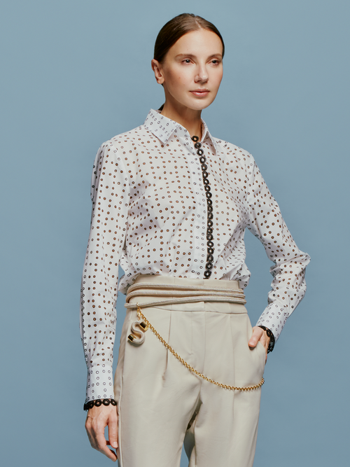 A woman stands against a blue background, wearing the white Naroa Blouse with black dots and light gray pants, accessorized with a chain belt, ready for the fresh styles of September 2nd, 2024.