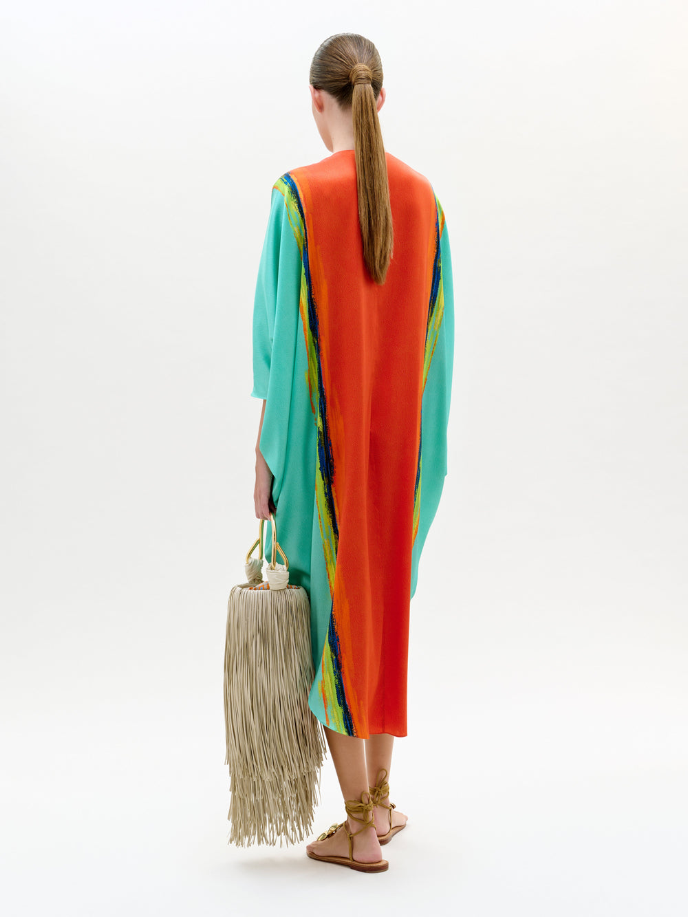 Woman wearing a colorful dress with a rainbow pattern, holding a beige bag with tassels on a white background.