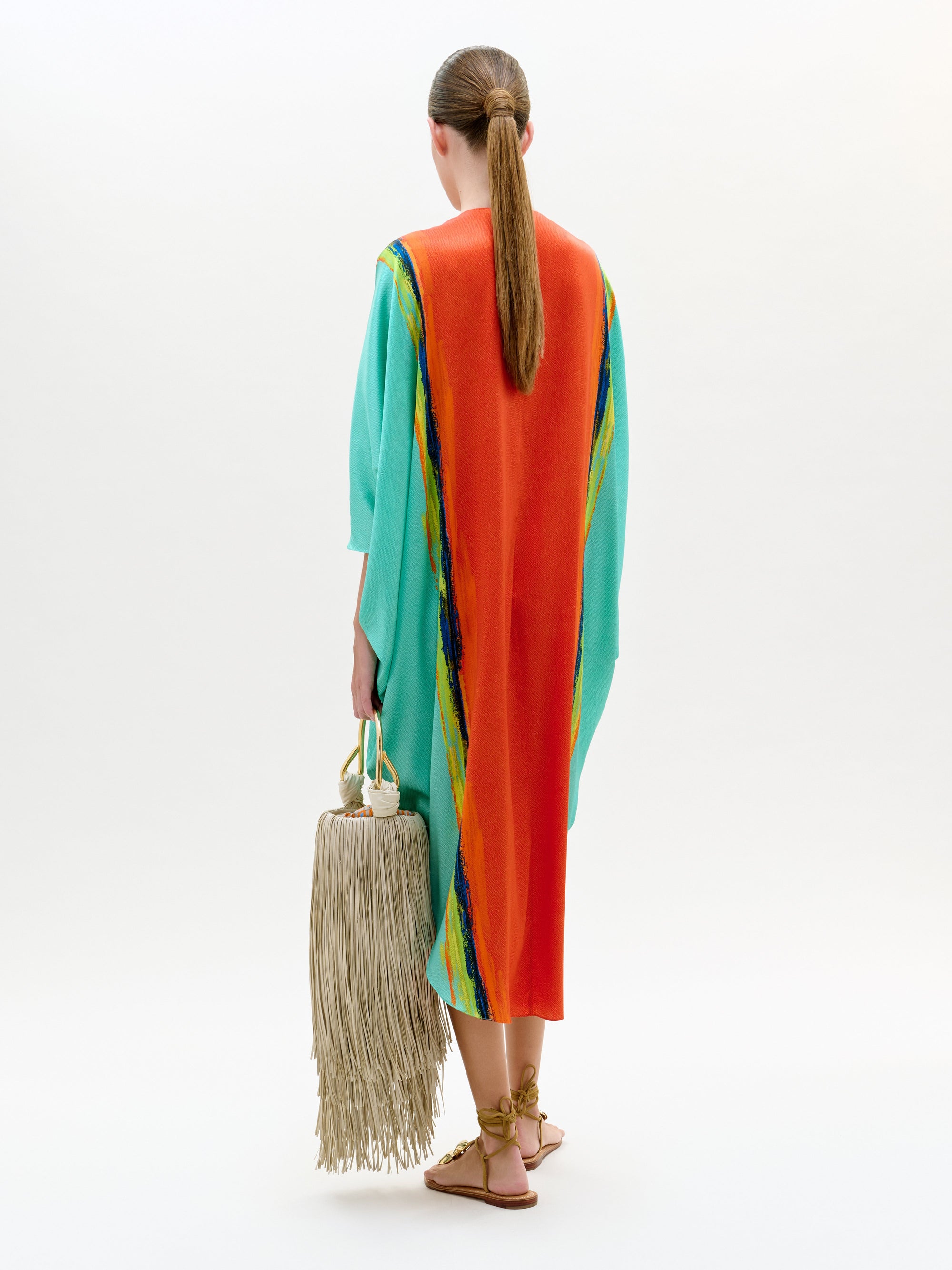 Woman wearing a colorful dress with a rainbow pattern, holding a beige bag with tassels on a white background.