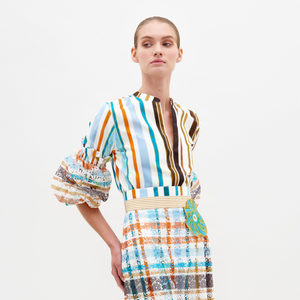 A woman stands against a plain background wearing the Nuru Blouse, a 100% cotton, multi-color striped top with puffed sleeves, paired with a patterned skirt. She looks slightly left with her hands behind her back.