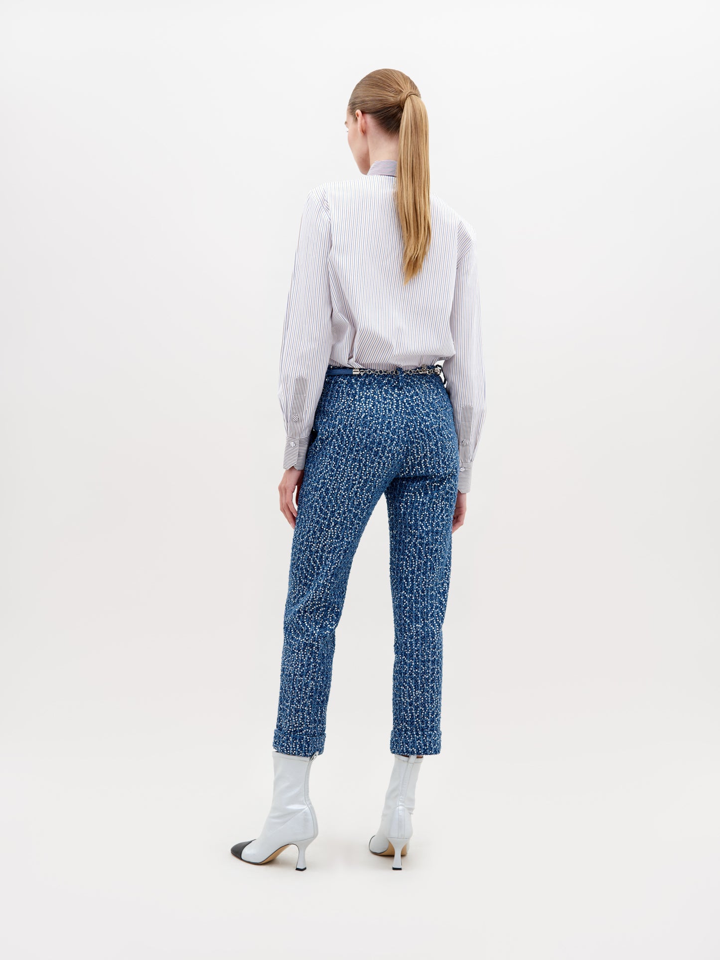 A woman stands against a plain background wearing a white blouse with blue lace detail, Garmet Pant Navy Sequins, a pearl belt, and white ankle boots with black tips. This stylish ensemble is available for pre-order now and will ship by November 15th, 2024.