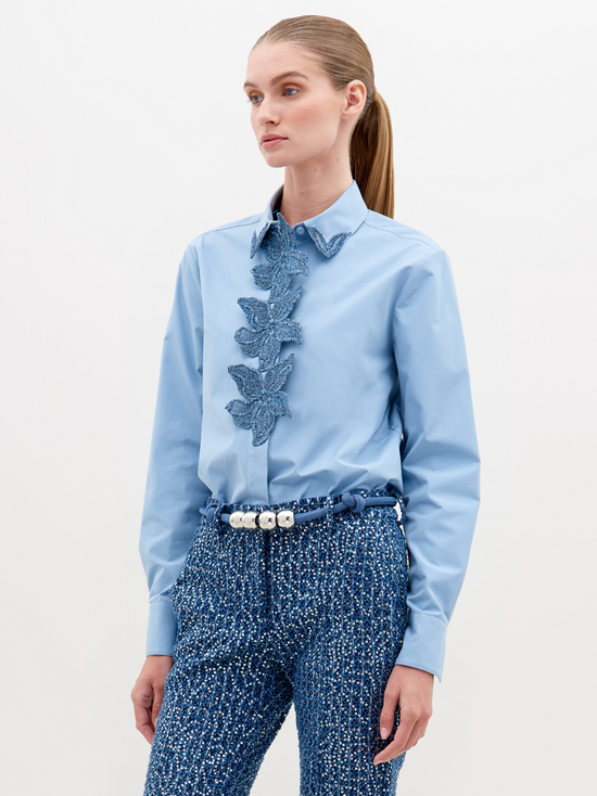 A woman with straight hair in a ponytail wears a Naroa Shirt Celestial Blue with ruffled detailing on the front and textured blue high-waisted pants, accessorized with a white beaded belt. Available for pre-order now, with an expected ship date in November 2024.