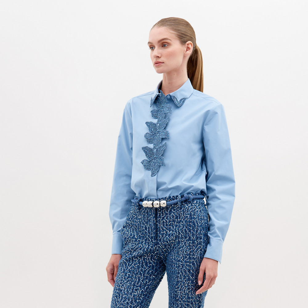 A woman with straight hair in a ponytail wears a Naroa Shirt Celestial Blue with ruffled detailing on the front and textured blue high-waisted pants, accessorized with a white beaded belt. Available for pre-order now, with an expected ship date in November 2024.