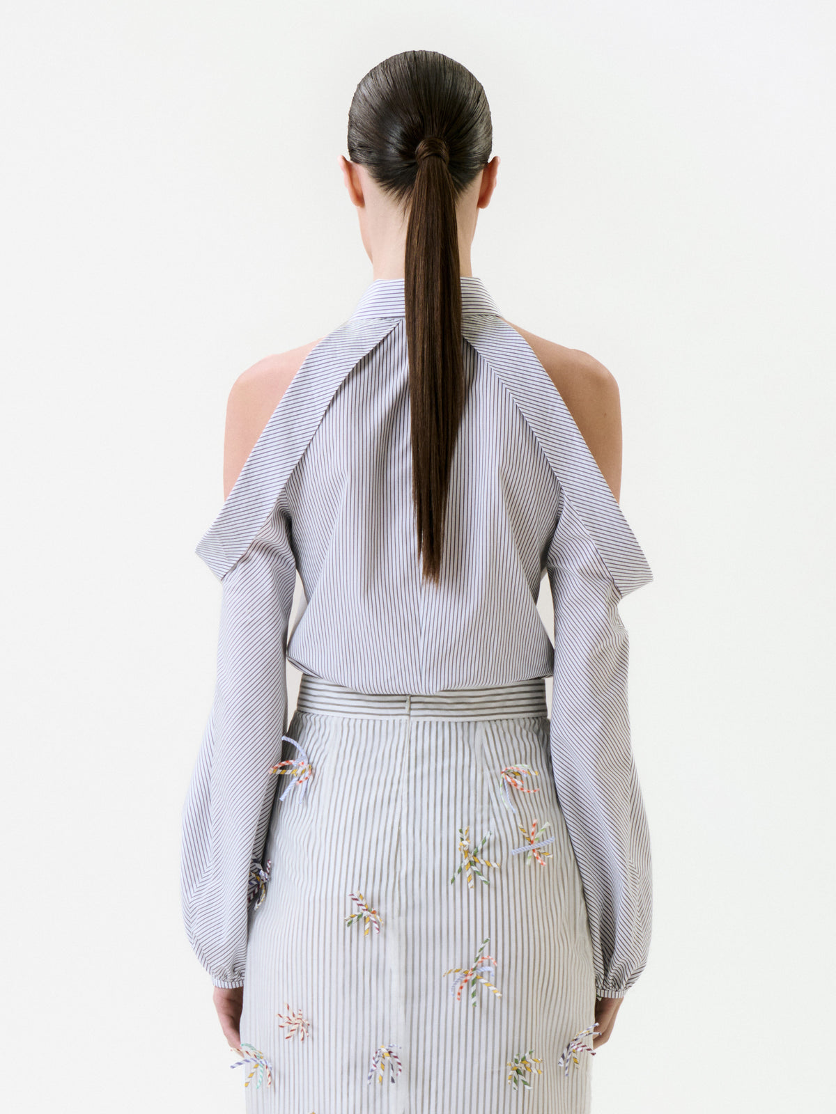 Back view of a person wearing a Nurith Shirt featuring light blue and white stripes with cold-shoulder cutouts and a matching striped skirt with small embroidered floral details.