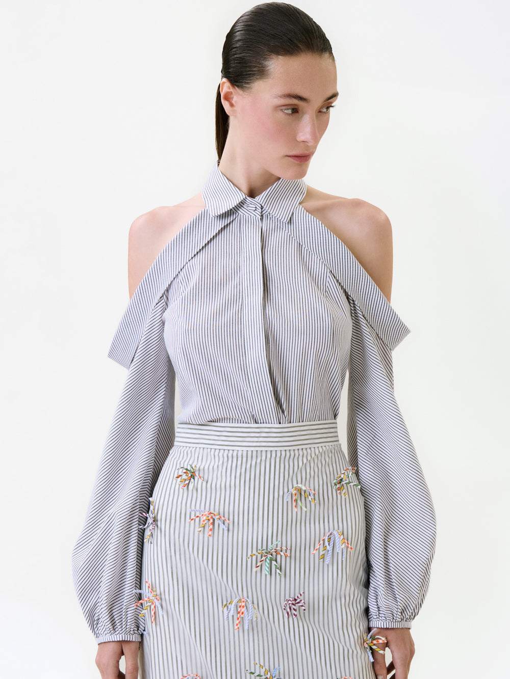 A model wearing a grey and white striped cold-shoulder Nurith Shirt and a matching high-waisted skirt adorned with small, colorful embroidered bows.