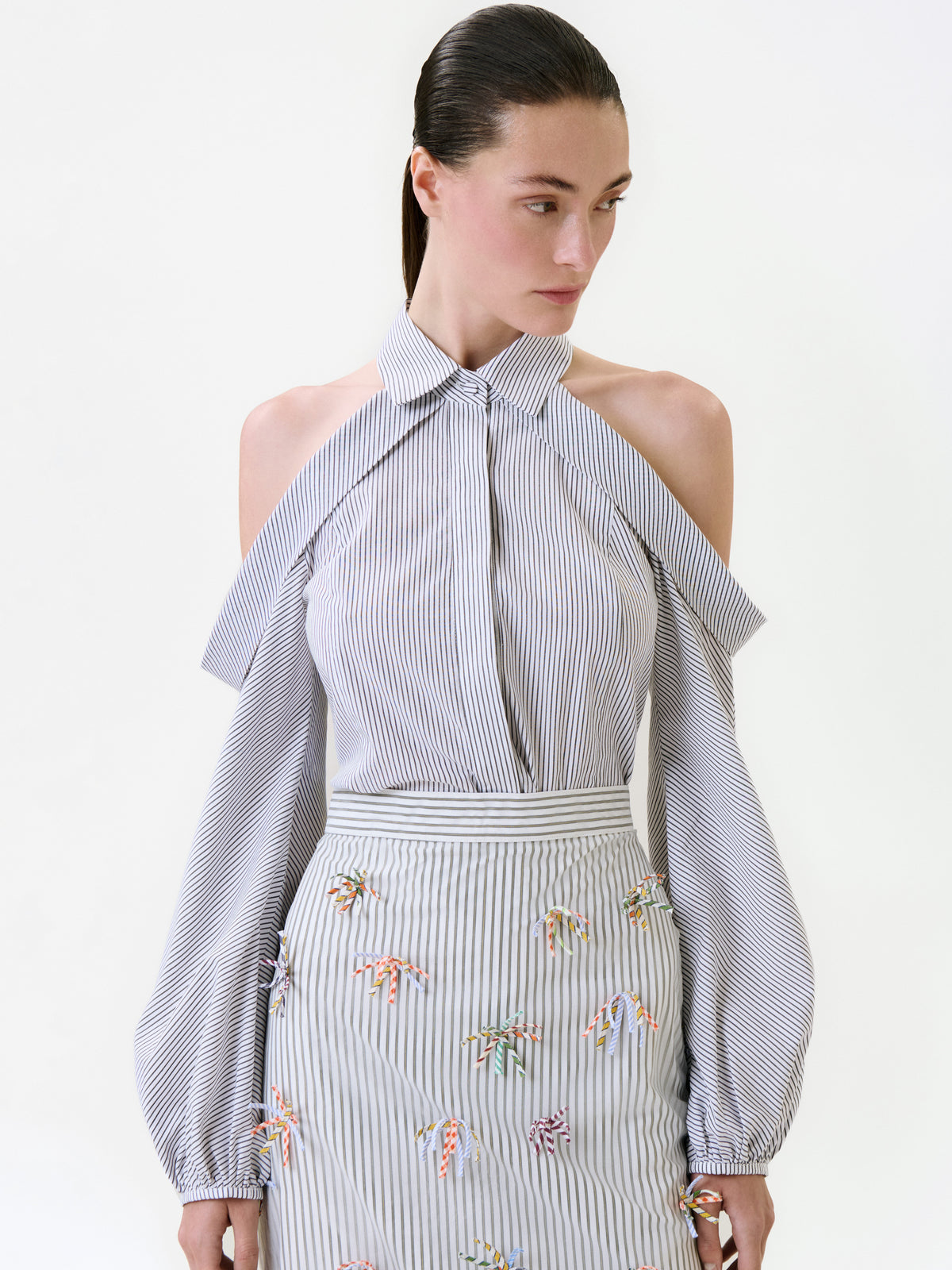 A model wearing a grey and white striped cold-shoulder Nurith Shirt and a matching high-waisted skirt adorned with small, colorful embroidered bows.