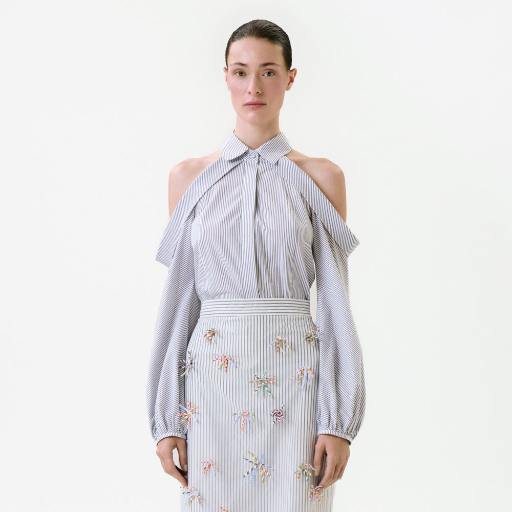 A person wears a grey and white pinstriped Nurith Shirt with cold-shoulder cutouts and long sleeves, tucked into a matching skirt adorned with small, colorful embroidered embellishments.