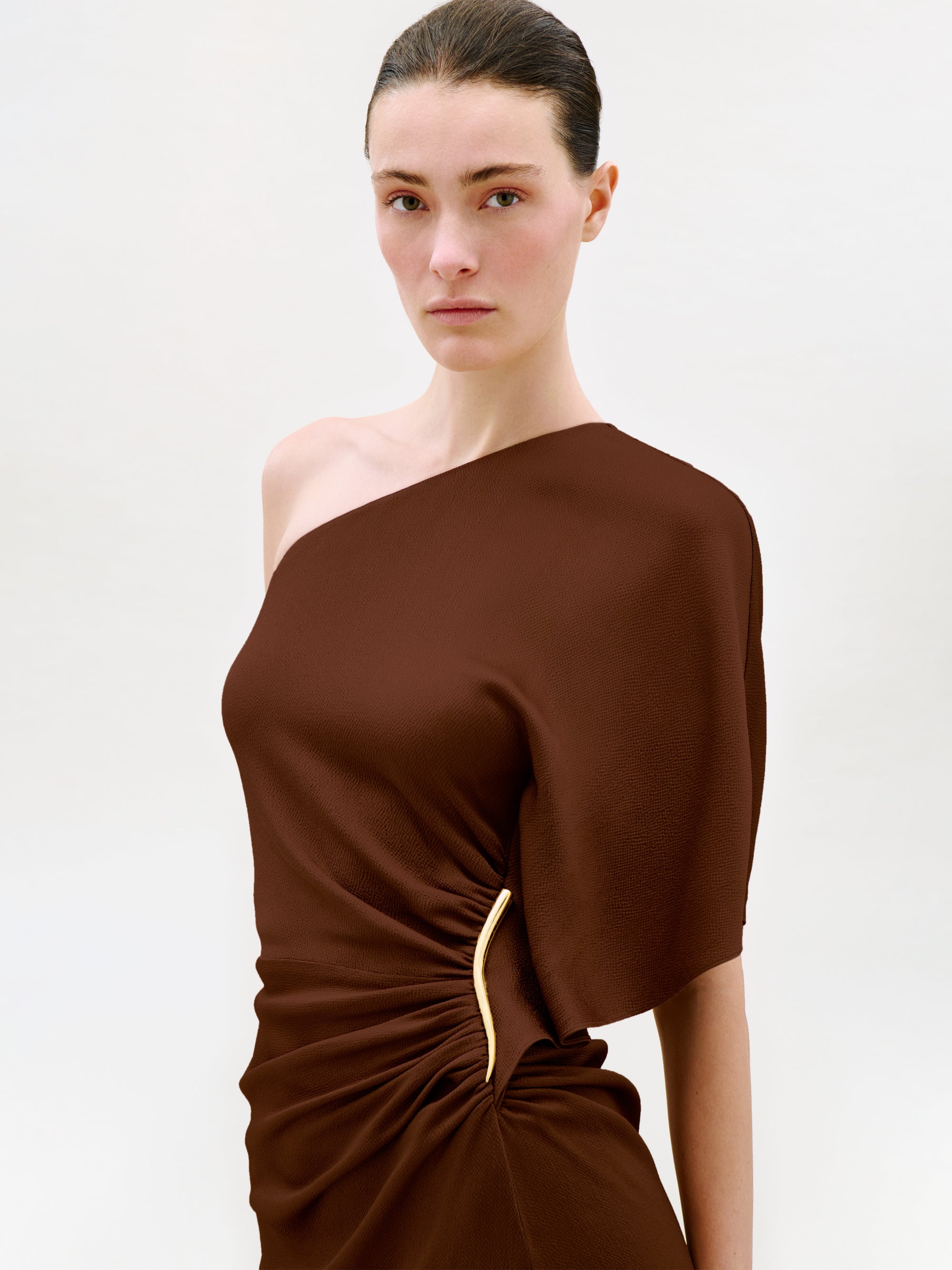 A woman with dark hair in a sleek bun wears the Oby Dress, a brown one-shoulder midi dress crafted from luxurious Italian fabric with a gold accent at the waist, standing against a plain light background.