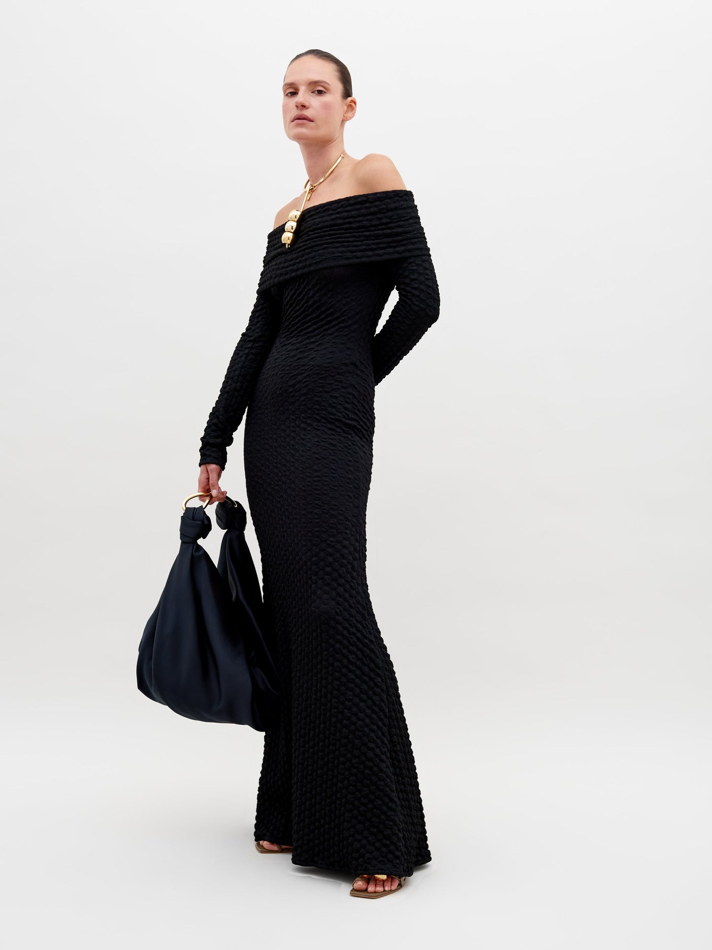 A person stands wearing a long, off-the-shoulder black textured Ozara Dress Black, holding a large black bag. They have a gold necklace and their hair is pulled back. The background is plain white, perfect for promoting the new collection available for pre-order. Mark your calendars for the ship by date: November 15th, 2024.