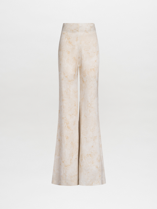 A woman stands wearing the Palermo Pant in Cream Abstract Print, a strapless jumpsuit featuring a light-colored fabric and a floral design on the top, accessorized with a small clutch against a plain blue background. Pre-order now for August shipping dates and elevate your summer wardrobe effortlessly.