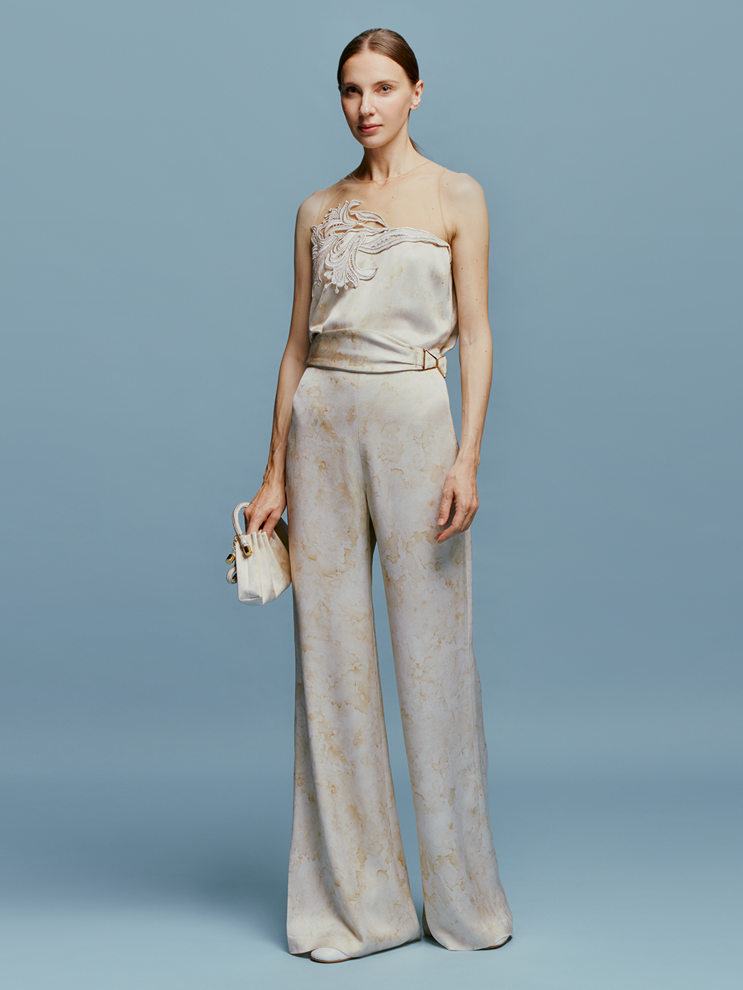 A woman stands wearing the Palermo Pant in Cream Abstract Print, a strapless jumpsuit featuring a light-colored fabric and a floral design on the top, accessorized with a small clutch against a plain blue background. Pre-order now for August shipping dates and elevate your summer wardrobe effortlessly.