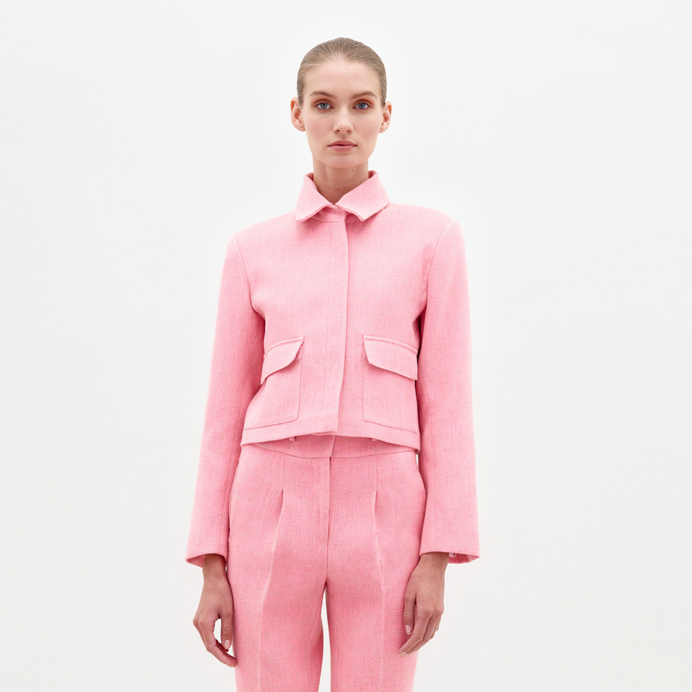 A person with tied-back hair is wearing a matching pink tailored suit, including the Perla Jacket Pink, which features a collar and two front pockets, standing against a plain white background. This stylish look will be available for pre-order, with the ship date set for February 2025.