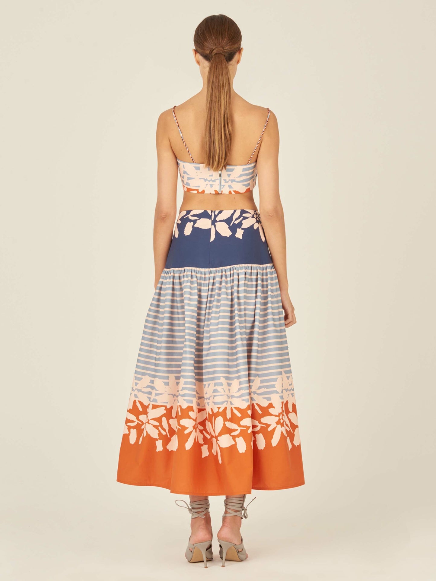 The Apia Skirt Celeste Orange is a flared midi design that features horizontal blue stripes and a wide blue and white floral band at the top. The hem is embellished with an eye-catching celeste orange floral border.