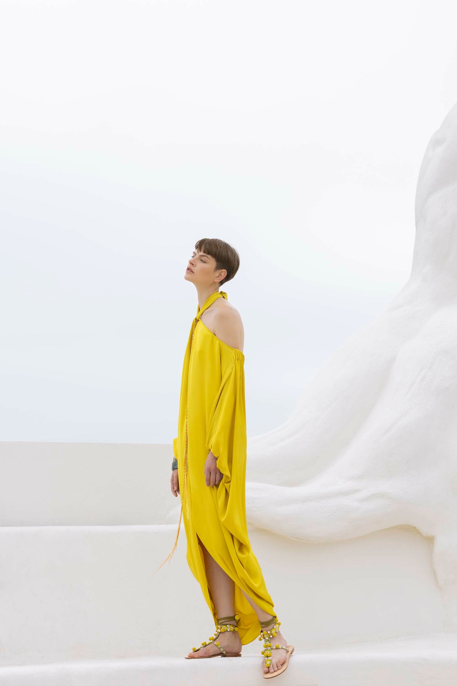 Person in a bright yellow off-shoulder dress stands against a large white abstract sculpture under a clear sky.