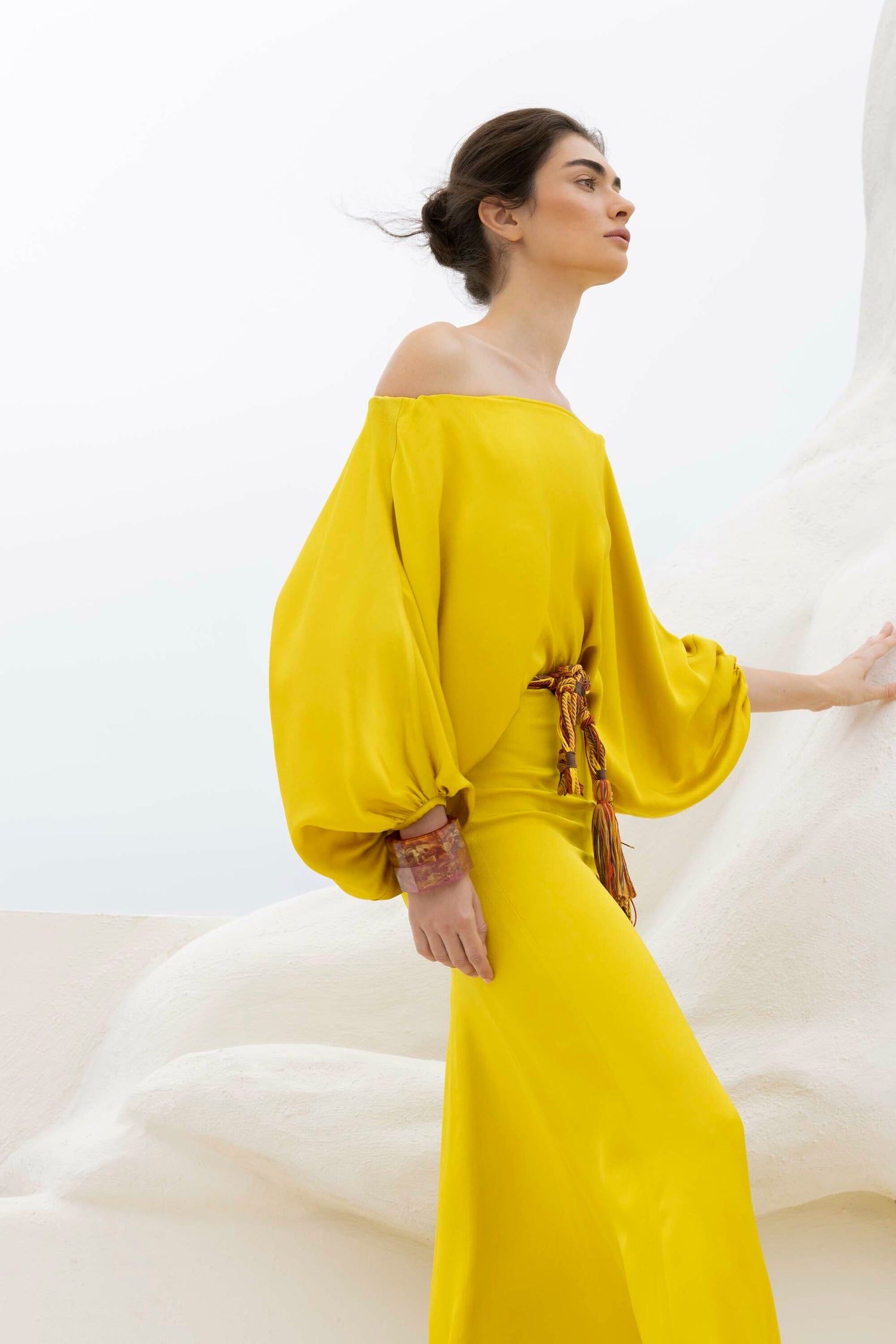 A person in a yellow off-shoulder dress with puffy sleeves stands by a large white sculpture, gazing to the side.