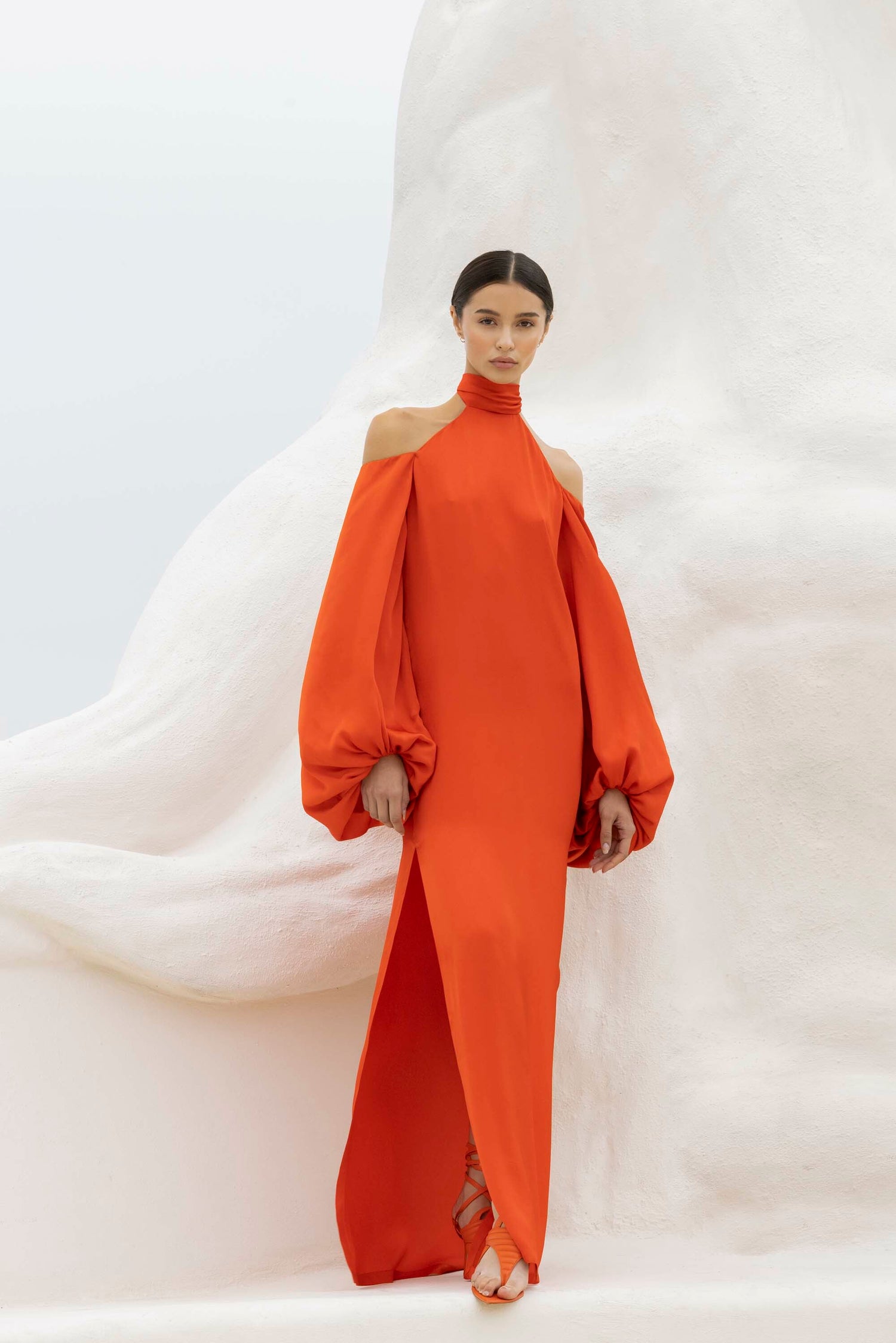 A person stands against a white abstract background wearing a long, orange dress with open shoulders and voluminous sleeves.