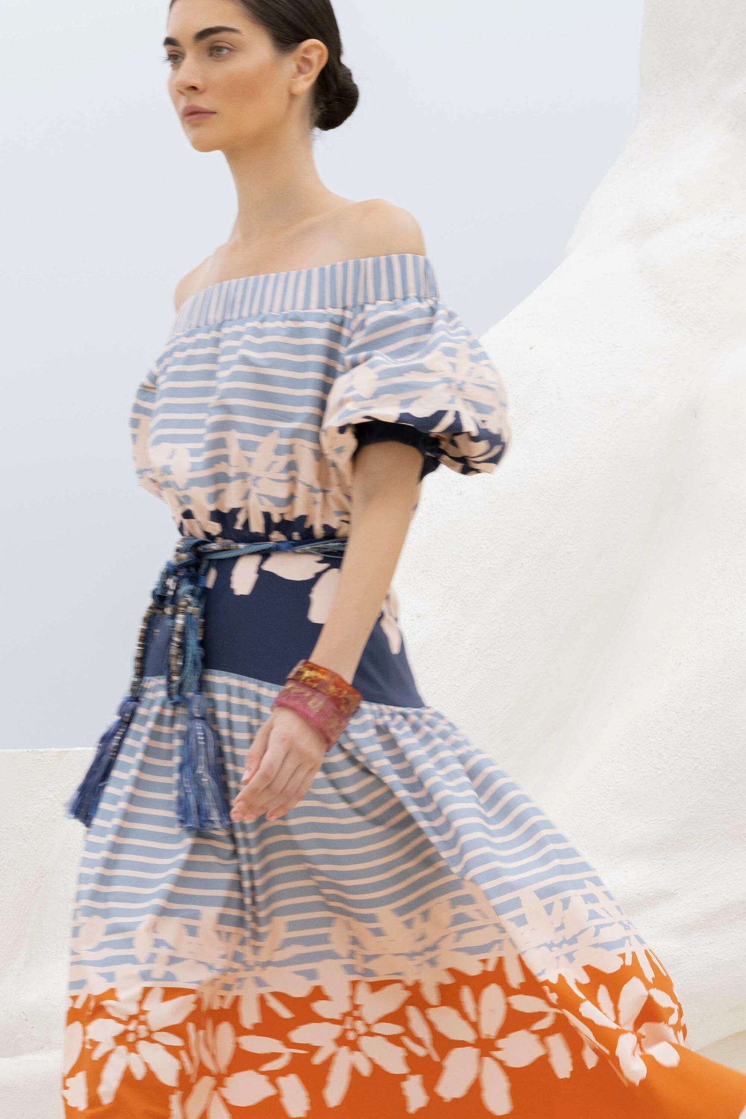 Person wearing a blue and white striped dress with floral patterns and a tasseled belt, walking against a light background.